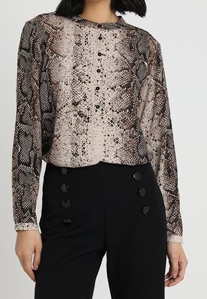 Kobieta nosząca bluzkę na guziki z długim rękawem w wężowy wzór oraz czarne spodnie o wysokim stanie z dwoma rzędami dużych guzików.