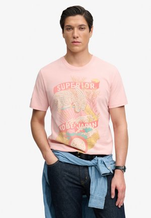 Uomo che indossa una T-shirt rosa con grafica di una gru e la scritta "Superior Kobe Japan", jeans blu, cintura nera, orologio e camicia blu annodata in vita.