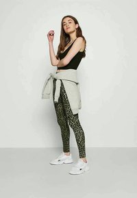 Leggings de estampa de leopardo verde, top preto, sweatshirt cinza atado e sapatilhas brancas; modelo posa com um fundo neutro.