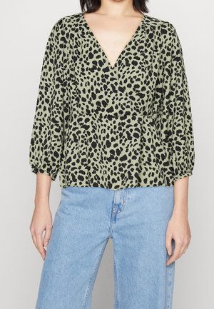 Woman wearing a green wrap blouse with black leopard spots and three-quarter sleeves paired with light blue high-waisted jeans.