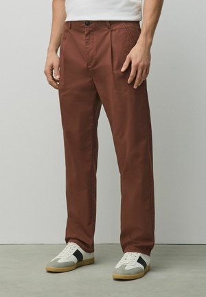 Man wearing brown straight-leg pants and white sneakers with grey and black accents, standing against a plain wall.