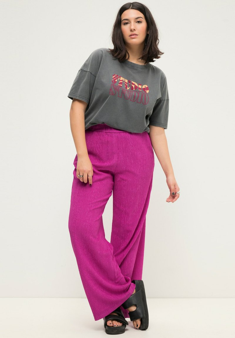 Grijze oversized t-shirt met rode grafische tekst, gecombineerd met wijde roze broeken. De broeken hebben een gestructureerde afwerking en een hoge taille. Zwarte platform sandalen.