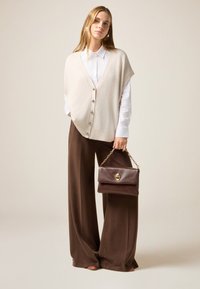 Chaleco de punto beige con cierre de botones sobre una camisa blanca, combinado con pantalones de pierna ancha marrones y un pequeño bolso marrón con cadena.