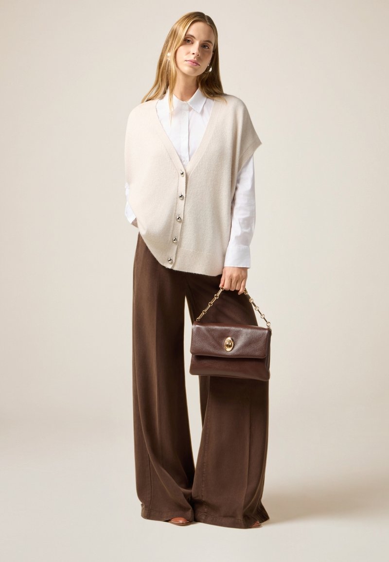 Chaleco de punto beige con cierre de botones sobre una camisa blanca, combinado con pantalones de pierna ancha marrones y un pequeño bolso marrón con cadena.