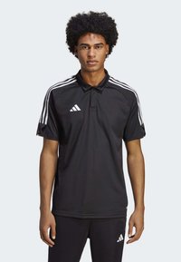 Polo shirt nero a maniche corte con colletto, caratterizzato da un design a tre strisce bianche sulle spalle e un logo Adidas sul lato sinistro del petto.