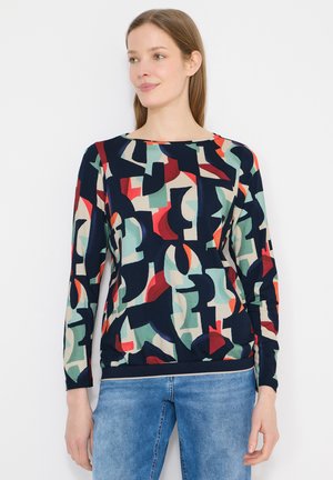 Femme portant un haut à manches longues avec un motif géométrique abstrait en bleu marine, rouge, vert et beige, associé à un jean bleu clair, debout contre un mur blanc.