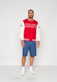 Rde varsity jakna z belimi rokavi, besedilom "CHIEFS" in ekipo logo na prsih. Nosi se z modrimi denim hlačami in belimi supergami.