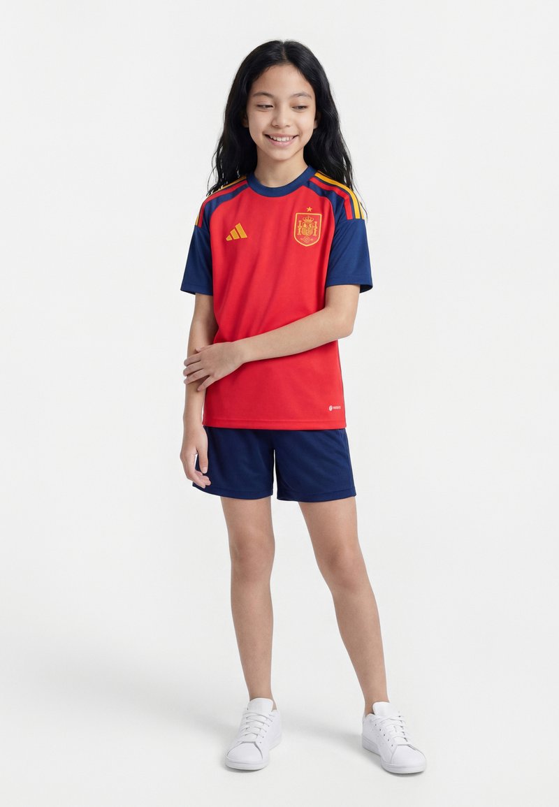 Jeune fille souriante, portant un maillot et un short de football espagnol rouge et bleu marine, debout avec un bras croisé sur l’autre, baskets blanches.