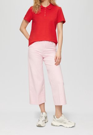 Nainen seisoo päällään punainen lyhythihainen poolopaita, vaaleanpunaiset leveälahkeiset lyhyet housut ja valkoiset paksupohjaiset lenkkarit.