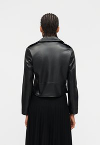 Άτομο με πλεξούδες που φοράει μαύρο δερμάτινο cropped μπουφάν και μαύρη πλισέ φούστα, εμφανιζόμενο από πίσω μπροστά σε απλό φόντο.