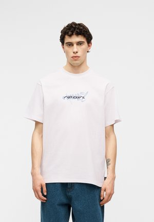 Ung mann med krøllete hår iført en lys rosa T-skjorte med Rip Curl-logo og blå jeans, stående foran en ensfarget hvit bakgrunn.