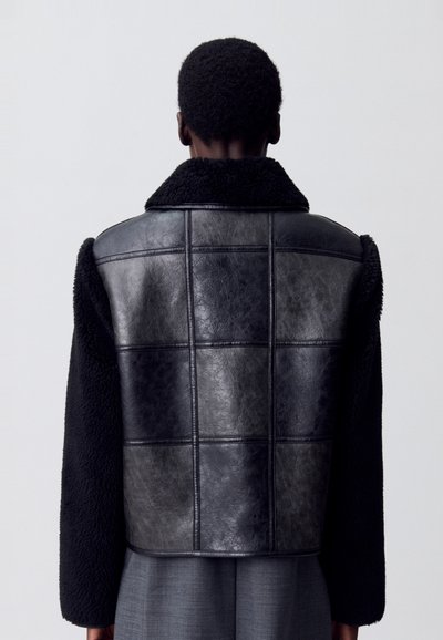Chaqueta negra de borreguito y cuero con un diseño de panel rectangular, que presenta un cuello, mangas texturizadas en negro y una forma estructurada.