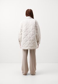 Jachetă din material bej deschis, matlasată, cu o croială lejeră, având modele în formă de diamant și gulere ascuțit. Asortată cu pantaloni largi din material bej deschis.