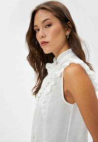 Blusa sin mangas blanca con cuello alto, que presenta acentos con volantes en la parte delantera. Textura suave, tejido ligero y detalles de botones.