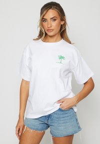 Mujer con cabello rubio largo viste una camiseta blanca con un gráfico de palmera verde y shorts de denim azul cortos, posando con una mano en el bolsillo.