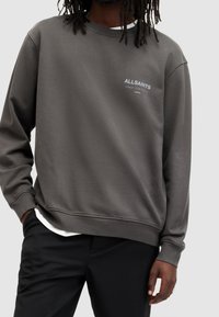 Grå sweatshirt i mjukt tyg med långa ärmar, rundad halsringning och en logotyp på bröstet. Buren över en vit skjorta.