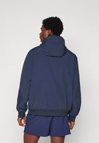 Navy blue bomber jacket made of lightweight fabric, featuring a hood, elastic cuffs, and hem, with a smooth texture and minimal detailing.