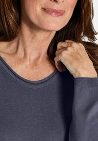 Frau mit braunem Haar, die einen grauen Strickpullover trägt, ihre Hand leicht an ihren Hals gelegt und mit einem schwachen Lächeln.