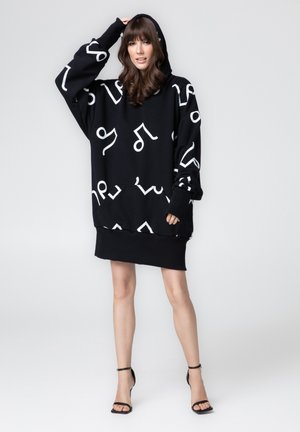 Kobieta ubrana w czarną, oversize'ową sukienkę dresową z białymi abstrakcyjnymi wzorami oraz czarne szpilki z paskiem wokół kostki, stojąca na jednolitym tle.
