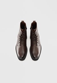 Barna bőr bokacsizma sima felülettel, elülső fűződizájnnal és oldalsó cipzárral rendelkezik, lekerekített orral és gumitalppal.