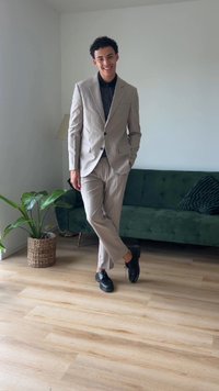 Light gray suit with slim-fit design and matching trousers, worn over a black shirt, paired with black shoes. Indoor setting with green sofa.