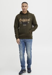 Olivovo zelená mikina s kapucňou a predným vreckom, s potlačou v vintage štýle. Nosená s svetlomodrými džínsami a čiernymi športovými topánkami.