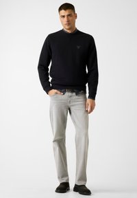 Fekete pulóver V-nyakú, logó részlettel. Világosszürke, egyenes szabású farmer és sötét cipők. Egységes, sima textúra.