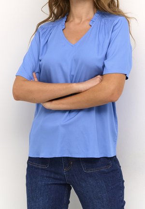 Femme portant un chemisier bleu clair à manches courtes en V avec des épaules à volants, bras croisés, associée à un jean en denim bleu foncé.