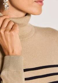 Nahaufnahme einer Person, die einen beigen Rollkragenpullover mit schwarzen Streifen anpasst und silberne geometrische Ohrringe trägt.