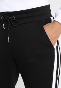 Svarta sweatpants med dragsko i midjan, sidofickor och kontrasterande vit rand längs den yttre sömmen.