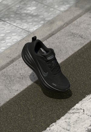 Czarny but do biegania Nike ze sznurowadłami, spoczywający na teksturowanym chodniku obok nawierzchni dotykowej i białego oznakowania drogi.