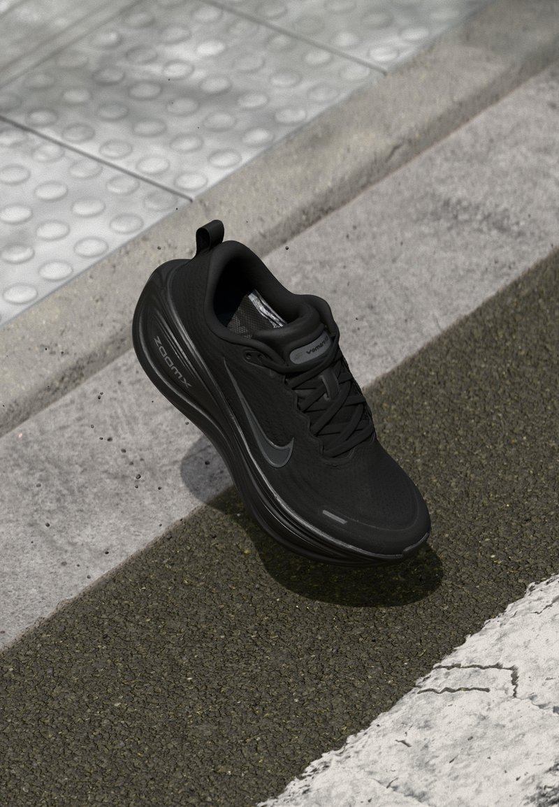 Chaussure de running noire Nike avec des lacets, posée sur un trottoir texturé près d’un revêtement tactile et d’un marquage au sol blanc.