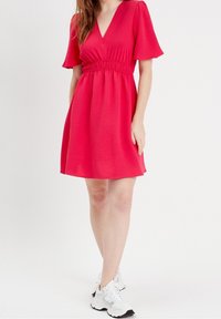 Robe courte rose avec un col en V, manches courtes évasées et taille cintrée. En tissu texturé, associée à des baskets blanches.