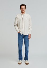 Männliches Modell steht, trägt ein beiges gestreiftes Hemd über einem weißen T-Shirt, blaue Jeans und beige Sneakers vor einem schlichten hellen Hintergrund.