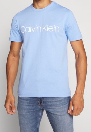 Mann trägt ein hellblaues Calvin Klein Kurzarm-T-Shirt kombiniert mit blauen Jeans, steht vor einem schlichten Hintergrund.