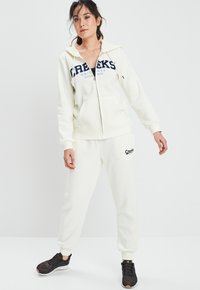 Helle Kapuzenjacke und Jogginghose Outfit, mit dem "Creeks" Logo in Marineblau, mit Reißverschluss und Taschenakzenten. Schwarze Sportschuhe runden den Look ab.