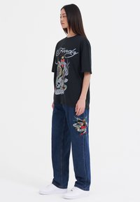 Black oversized t-shirt with colorful skull and snake graphic; paired with blue jeans featuring embroidered designs, worn with white sneakers.