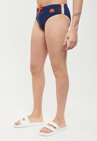Costume da bagno blu navy con cordino arancione e logo, caratterizzato da strisce orizzontali azzurre e bianche sui lati. Materiale liscio e elasticizzato.