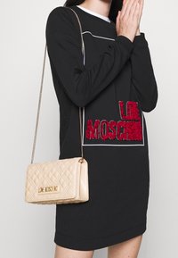Black long-sleeve dress with white accents and red embroidered logo; a beige quilted crossbody bag with a gold hardware plaque.