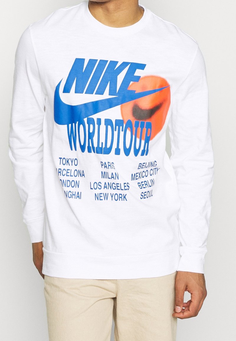 Valkoinen pitkähihainen huppari, jossa on sininen ja oranssi "NIKE WORLD TOUR" -grafiikka. Tekstissä on sinisellä painettuna kaupunkien nimet etupuolella.