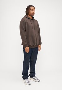 Brun hettegenser med frontlomme, kombinert med mørkeblå jeans og grå sneakers med hvite detaljer. Avslappet passform, myk tekstur.