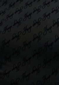 Tissu noir avec un motif diagonal répétitif du mot « Hugo » en écriture cursive.