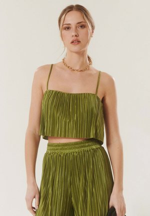 Jeune femme portant un crop top plissé vert et un pantalon plissé taille haute assorti, accessoirisé d'un collier chaîne dorée épaisse.