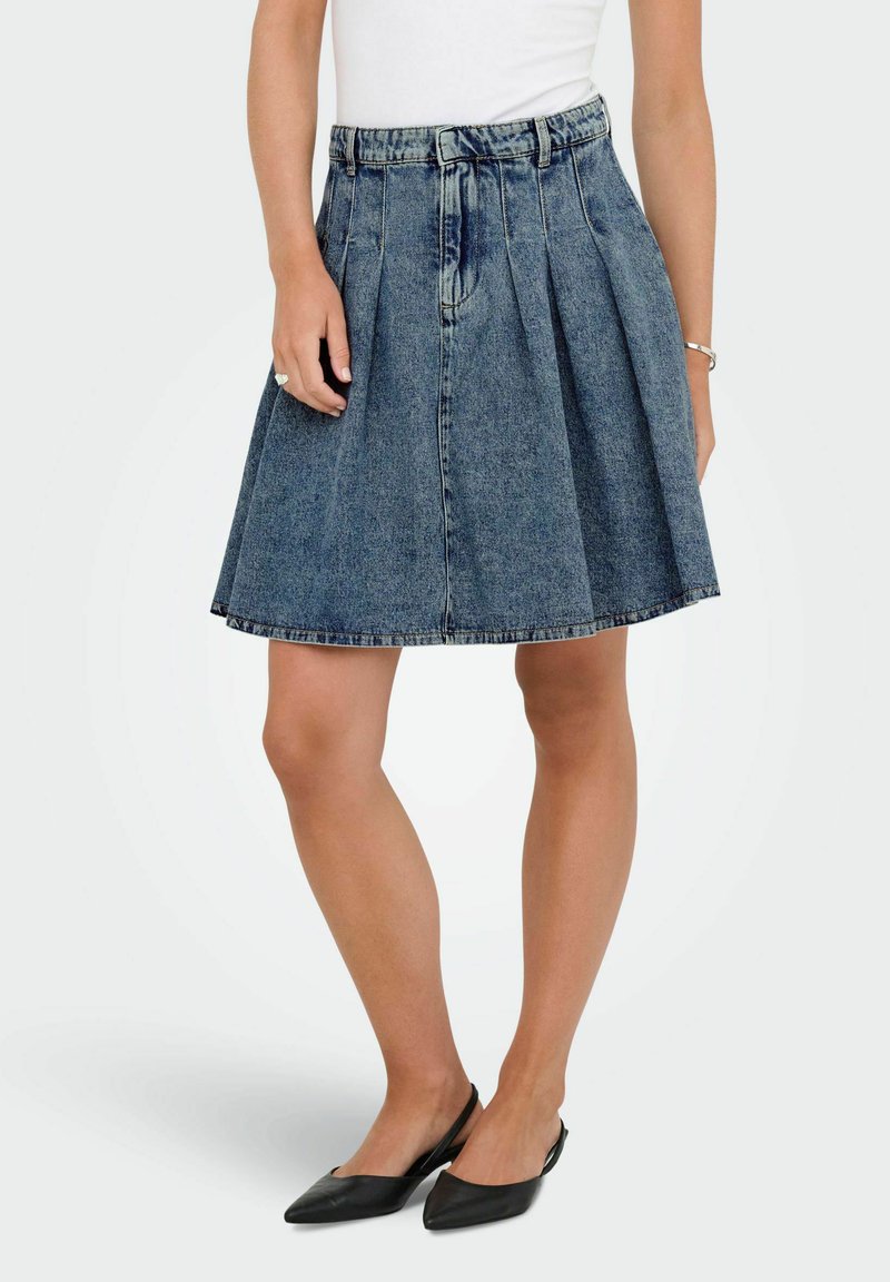 Jupe en denim bleu en forme A, avec des plis, une coupe mi-longue et une ceinture. Assortie à des chaussures noires à talons ouverts.