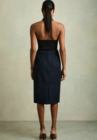 Mujer con el pelo largo y oscuro recogido en una coleta, que lleva una blusa negra sin tirantes, falda midi azul marina y tacones altos beige, de pie frente a una pared lisa.