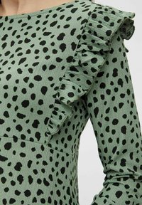 Zelené šaty s černým skvrnitým vzorem, strukturovanou tkaninou a volánovými akcenty na ramene. Dlouhé rukávy a přiléhavý střih.