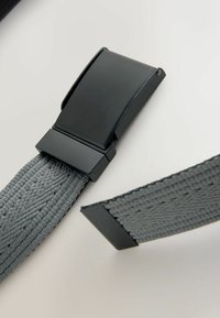 Boucle métallique noire avec sangle en tissu gris tissé, montrant des parties séparées prêtes à être reliées sur une surface claire.