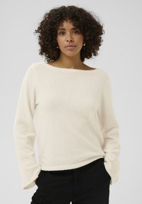 Pull crème duveteux avec un large décolleté bateau et des manches longues, présentant une coupe décontractée et une texture douce. Associé à un pantalon noir.
