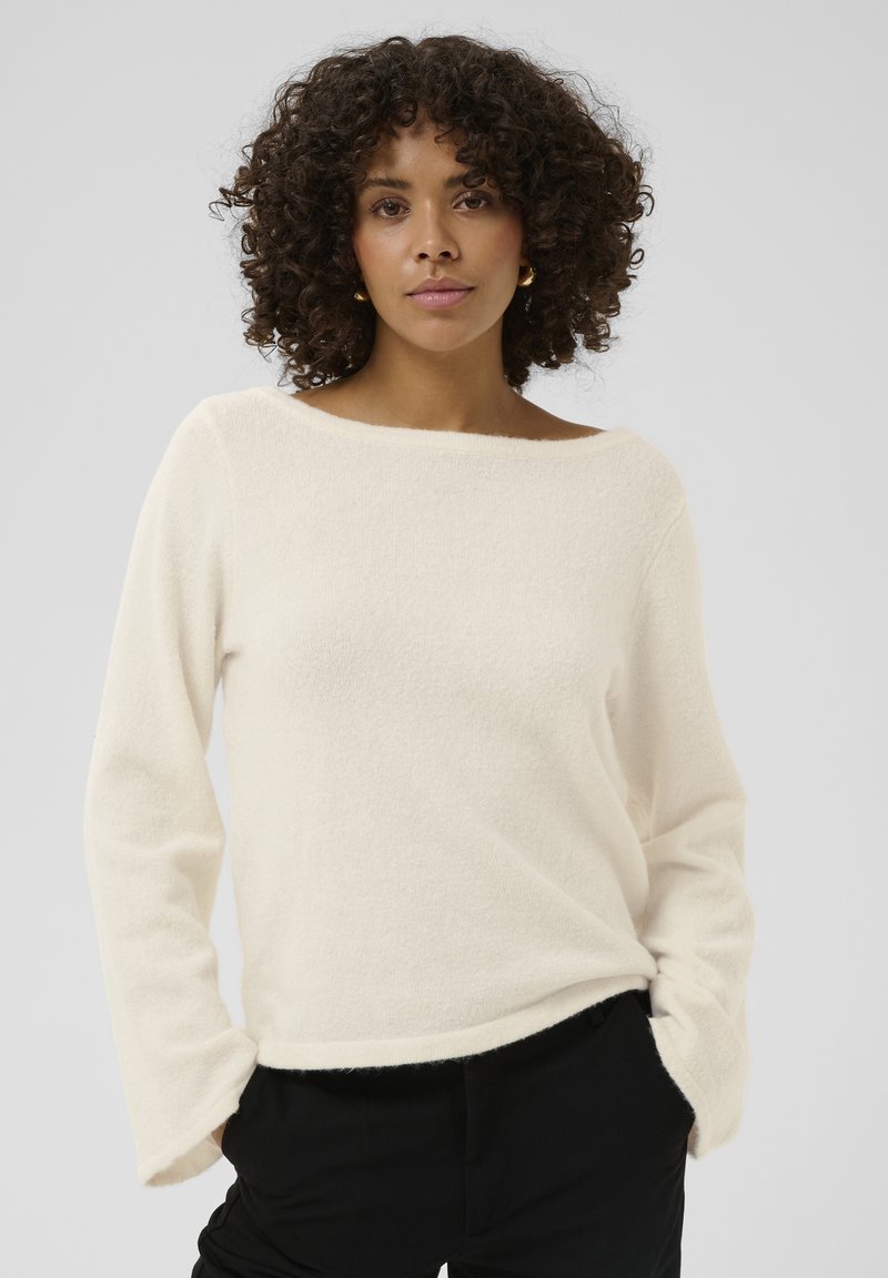 Pull crème duveteux avec un large décolleté bateau et des manches longues, présentant une coupe décontractée et une texture douce. Associé à un pantalon noir.