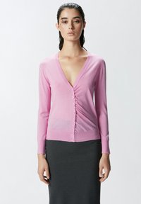 Svijetloružičasti kardigan s dubokim V-izrezom, dugim rukavima i prednjim gumbima. Mekani pleteni materijal s uskim krojem, upareno s tamnosivom suknjom.